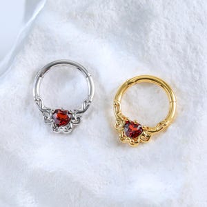 以下が含まれることがあります： 2つのセプタムリング。1つはシルバー、もう1つはゴールドで、それぞれハート型の赤い宝石が付いています。リングには宝石の周りに装飾的なデザインがあります。ジュエリーは白いテクスチャのある表面に展示されています。