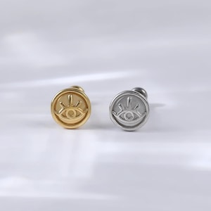 May include: Two small, round earrings with an eye design. The earring on the left is gold and the earring on the right is silver.