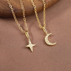 May include: Two gold-colored necklaces with delicate chains. One pendant is a four-pointed star, the other a crescent moon. Both pendants are embellished with small, sparkling details. The necklaces rest on a wooden surface.