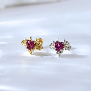 Può includere: Due piccoli orecchini a forma di cuore con ali di pipistrello, uno in oro e uno in argento, con una gemma rosa al centro.