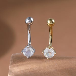 May include: Two belly button rings. One is silver-colored, the other is gold-colored. Each has a small, round ball at the top and a clear, teardrop-shaped gem at the bottom. The background is a solid, warm brown.