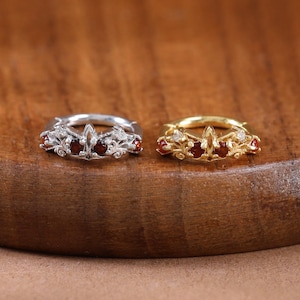 Könnte beinhalten: Zwei kleine Creolen, eine aus silberfarbenem und eine aus goldfarbenem Metall, die jeweils ein filigranes Design mit kleinen, dunkelroten Steinen aufweisen, die in den vorderen Teil der kreisförmigen Ringstruktur eingelassen sind.