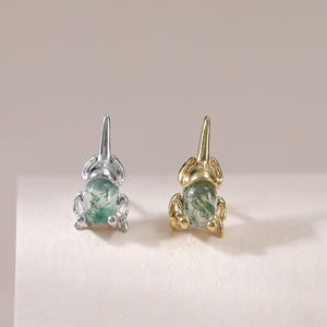 May include: Two stud earrings shaped like small frogs, each featuring an oval moss agate gemstone as the body. One earring is crafted from silver-toned metal, while the other is made from gold-toned metal, both showcasing detailed limbs.