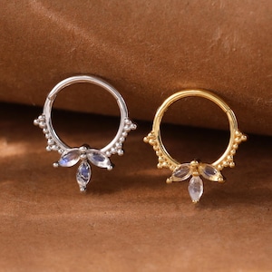 May include: Two septum rings, one silver and one gold, each with a decorative design. The silver ring has a cluster of three teardrop-shaped stones and small beaded accents. The gold ring has a similar design. Both are set against a brown background.