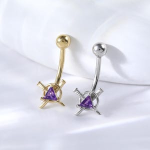 May include: Two belly button rings with a purple triangle and crossed swords design. One ring is gold and the other is silver.