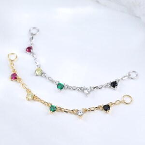 Könnte beinhalten: Zwei filigrane Ketten-Schmuckstücke, eines in Gold und eines in Silber, jeweils mit farbigen Edelsteinen verziert. Die goldene Kette hat rote, gelbe, grüne und schwarze Steine. Die silberne Kette hat rote, grüne, weiße und schwarze Steine. Der Schmuck wird auf einer weißen Oberfläche präsentiert.