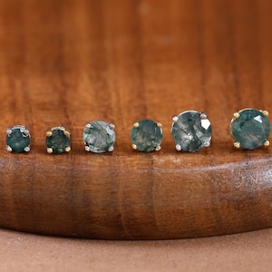 May include: A set of six round stud earrings featuring moss agate gemstones in gold-toned settings. The stones display varying shades of deep green with natural inclusions, arranged in a line from smallest to largest on a curved wooden surface.