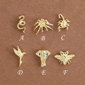 Könnte beinhalten: Sechs kleine goldfarbene Metallanhänger in zwei Reihen, beschriftet von A bis F. Die Anhänger zeigen eine Schlange, eine Spinne, einen Skorpion, einen Kolibri, einen Elefantenkopf und eine Biene. Jedes Stück hat detaillierte Texturen.