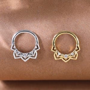 May include: Two septum rings, one silver and one gold, each with a floral design. The silver ring has a polished finish, while the gold ring has a warm, radiant hue. Both rings feature small, clear gemstones.