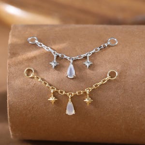 Puede incluir: Dos delicados collares de cadena, uno plateado y otro dorado, cada uno adornado con abalorios de estrella y un colgante en forma de lágrima. Los collares se exhiben sobre una superficie marrón, mostrando su intrincado diseño.