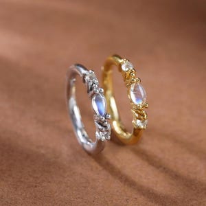 Könnte beinhalten: Zwei Creolen, eine in Silberoptik und eine in Goldoptik, jeweils mit einem zentralen, irisierenden Stein im Marquise-Schliff, flankiert von kleinen, klaren, runden Steinen. Die Ringe stehen aufrecht und zeigen ihre feinen Metallbänder.