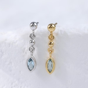 Può includere: Due orecchini pendenti, uno argentato e uno dorato, ciascuno con una serie di piccole sfere che conducono a una gemma blu chiaro a forma di marquise. Gli orecchini sono esposti su uno sfondo bianco strutturato.