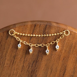 May include: A delicate gold necklace with a double-layered design. The top layer is a beaded chain, and the bottom layer features a chain with four small, dangling clear gemstones. The necklace is displayed on a wooden surface.