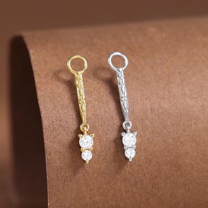 Puede incluir: Dos pendientes colgantes, uno dorado y otro plateado, cada uno con un pequeño lazo en la parte superior y una barra vertical decorativa. La parte inferior de cada pendiente tiene dos piedras preciosas redondas y transparentes. Los pendientes se muestran sobre una superficie marrón.