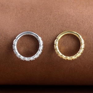 May include: Two hoop earrings, one silver and one gold, are displayed against a brown backdrop. Both earrings have a textured surface. The silver earring is on the left, and the gold earring is on the right.