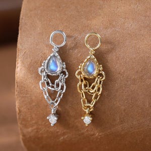 May include: Two ornate earrings, one silver-toned and one gold-toned, each featuring a teardrop-shaped, iridescent gemstone. The earrings have chain and filigree details, with a small, clear gemstone at the bottom.