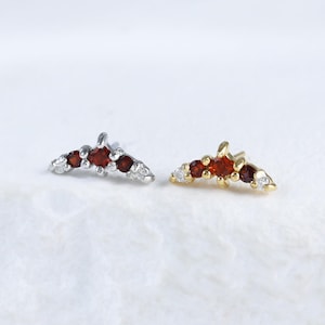 May include: Two stud earrings, one silver and one gold, each featuring a curved design. The silver earring has five red gemstones. The gold earring has three red gemstones and two white gemstones. Both earrings are on a white surface.