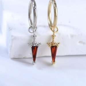 May include: Two hoop earrings, one silver-toned and one gold-toned, each featuring a dangling pendant shaped like a small sword or dagger. The pendants are accented with a triangular, deep red gemstone set into the metal base of the blade.