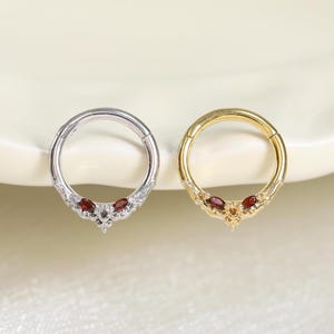 Peut inclure: Deux anneaux segmentés articulés circulaires, l'un argenté et l'autre doré. Chaque anneau présente un motif en arc pointu décoratif sur la partie inférieure, orné de petites pierres claires et de deux pierres rouge foncé taille marquise.