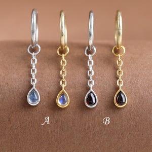 Puede incluir: Cuatro pendientes de aro con cadenas colgantes y piedras en forma de lágrima. Dos son plateados con piedras azules y dos son dorados con una piedra azul y otra roja oscura. Las letras A y B aparecen impresas bajo el segundo y cuarto pendiente.