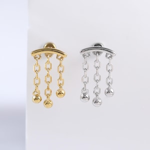 Puede incluir: Dos pendientes, uno dorado y otro plateado, cada uno con una barra curva y cadenas colgantes con pequeñas esferas. Se presentan sobre un fondo blanco, resaltando su diseño y detalles.