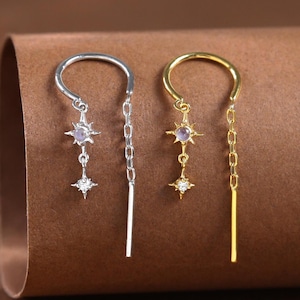 Może przedstawiać: Dwa kolczyki o tematyce niebiańskiej. Jeden jest w kolorze srebrnym, drugi w kolorze złotym. Każdy kolczyk posiada gwiazdę i zwisający łańcuszek z pręcikiem na końcu. Kolczyki mają górną część w kształcie półksiężyca, która zaczepia się w uchu.