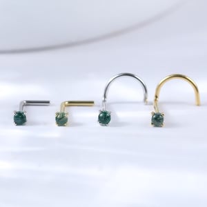 May include: Four nose studs, each featuring a small, round, green gemstone. The studs are in various styles: a silver L-shape, a gold L-shape, a silver screw, and a gold screw. The gemstones are set in a minimalist design.