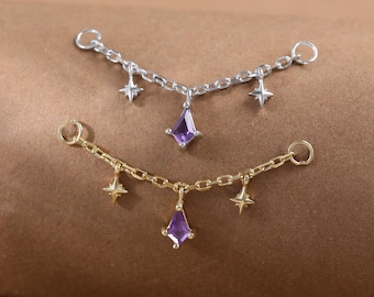 May include: Two delicate jewelry pieces, one silver and one gold, each featuring a chain with star charms and a central purple gemstone pendant. The jewelry is displayed on a brown surface.