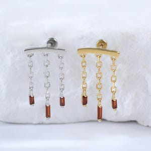 May include: Two earrings, one silver and one gold, each with a horizontal bar and three dangling chains. Each chain has a small, rectangular, reddish-brown bead at the end. The earrings are displayed on a white, textured surface.