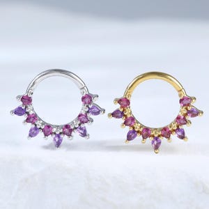 Puede incluir: Dos pendientes de aro, uno plateado y otro dorado, cada uno adornado con un patrón de rayos de sol de piedras preciosas moradas y rosas. Las piedras preciosas en forma de lágrima están dispuestas alrededor de los aros, creando un efecto brillante.