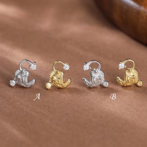 Pode incluir: Quatro brincos em forma de peixe-pescador dispostos em fila, incluindo duas peças prateadas e duas douradas. Cada brinco possui uma pequena pedra transparente na isca, com as letras A e B posicionadas abaixo dos pares.