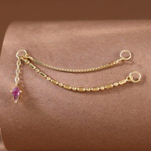 Puede incluir: Una cadena conectora de joyería en tono dorado que presenta dos estilos de cadena distintos, uno con eslabones cuadrados lisos y otro con cuentas cilíndricas alternas, terminado con un dije de piedra rosa en forma de cometa y dos anillas.