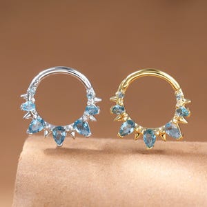 May include: Two hoop earrings, one silver and one gold, each featuring light blue teardrop gemstones. The earrings are displayed side-by-side against a brown backdrop, highlighting their design and colour contrast. The gemstones are evenly spaced around the hoops.