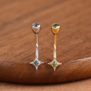 May include: Two curved eyebrow rings. One is silver-coloured, the other gold-coloured. Each has a small, round, green gemstone at the top and a four-pointed star with a green gemstone at the bottom. The background is a wooden surface.