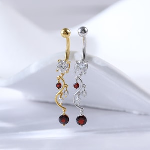 May include: Two belly button rings with a dangling design. One ring is gold with a red gemstone and the other is silver with a red gemstone.