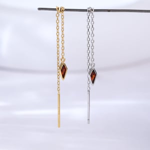 Op de afbeelding: Een paar oorhangers, één goudkleurig en één zilverkleurig, elk met een kleine, rode, ruitvormige edelsteen hanger. De oorbellen hangen aan een fijne ketting en hebben een verticale staaf aan de onderkant.