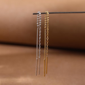 以下が含まれることがあります： 暗い棒から吊り下げられた、銀色と金色の一対のぶら下がりピアス。 各イヤリングは、端にまっすぐなバーが付いた繊細なチェーンが特徴です。 背景は無地の温かみのある茶色です。