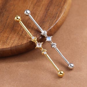 May include: Two industrial bar earrings, one gold and one silver, each featuring celestial-themed charms. The gold earring has a star, moon, and round ball accents. The silver earring has a star, moon, and a small square stone.