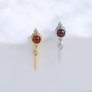 Peut inclure: Deux boucles d'oreilles, l'une dorée et l'autre argentée, chacune ornée d'une petite pierre précieuse ronde brun rougeâtre. Les boucles d'oreilles ont un dessus décoratif et un charm rectangulaire pendant. Elles sont présentées sur une surface blanche.