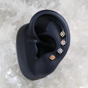 May include: Four small square earrings with a gold setting and colorful gemstones. The earrings are displayed on a black ear model.