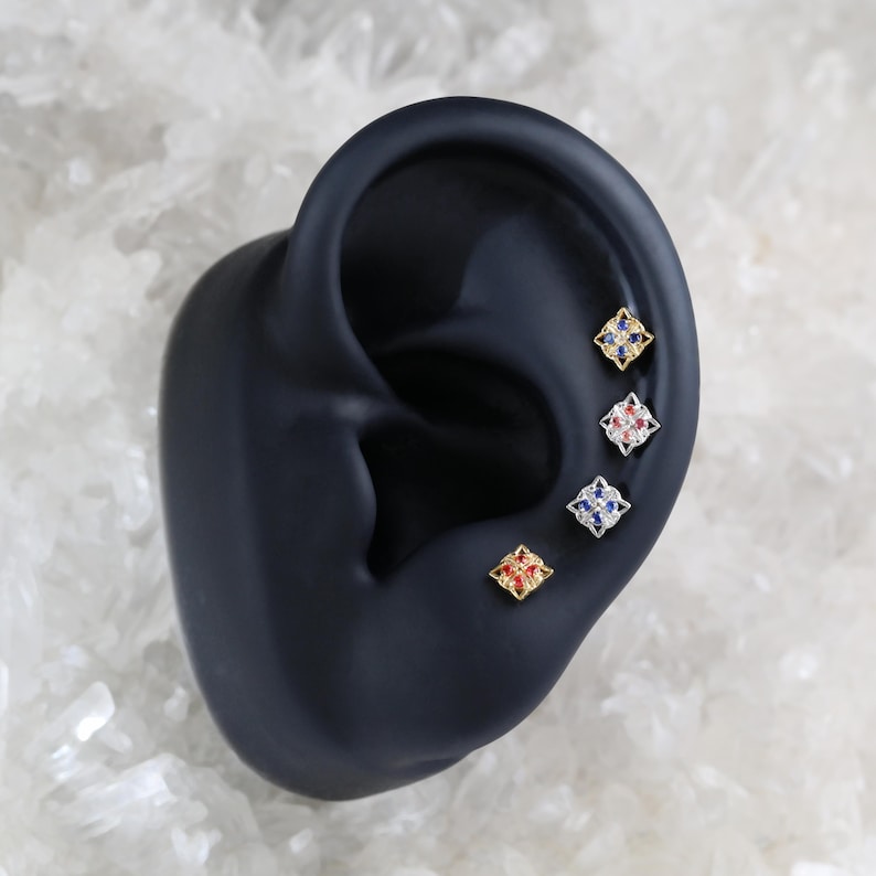 May include: Four small square earrings with a gold setting and colorful gemstones. The earrings are displayed on a black ear model.