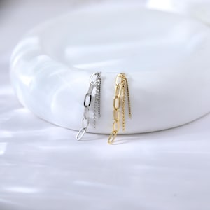 May include: A pair of chain earrings, one silver and one gold, displayed on a white surface. The earrings feature a chain design with dangling strands of smaller chains. The gold earring has a polished finish, while the silver earring has a similar design.