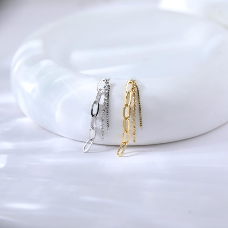 May include: A pair of chain earrings, one silver and one gold, displayed on a white surface. The earrings feature a chain design with dangling strands of smaller chains. The gold earring has a polished finish, while the silver earring has a similar design.