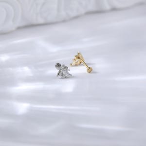 Puede incluir: Dos pendientes de bot&oacute;n sobre una superficie blanca. Uno es plateado con un dise&ntilde;o de hoja y una gema transparente. El otro es dorado con un dise&ntilde;o de rayos de sol. Ambos tienen un poste y un respaldo para orejas perforadas.