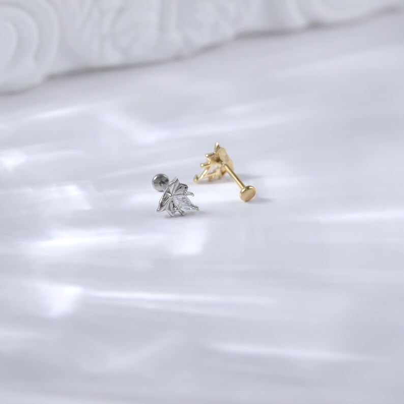 Puede incluir: Dos pendientes de bot&oacute;n sobre una superficie blanca. Uno es plateado con un dise&ntilde;o de hoja y una gema transparente. El otro es dorado con un dise&ntilde;o de rayos de sol. Ambos tienen un poste y un respaldo para orejas perforadas.