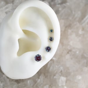 May include: A close-up of a white ear model with four round, blue-purple gemstones set in silver studs. The gemstones are different sizes, with the largest at the bottom and the smallest at the top.
