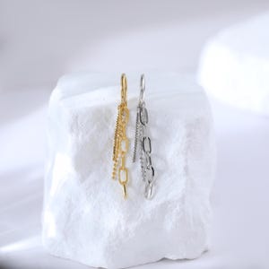 May include: A pair of dangling earrings, one gold-toned and one silver-toned, each featuring a chain design. The earrings hang from a small hoop and include multiple chain styles, including a delicate beaded chain.