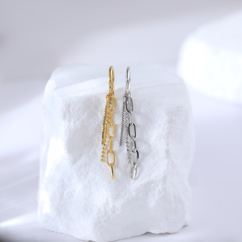 May include: A pair of dangling earrings, one gold-toned and one silver-toned, each featuring a chain design. The earrings hang from a small hoop and include multiple chain styles, including a delicate beaded chain.