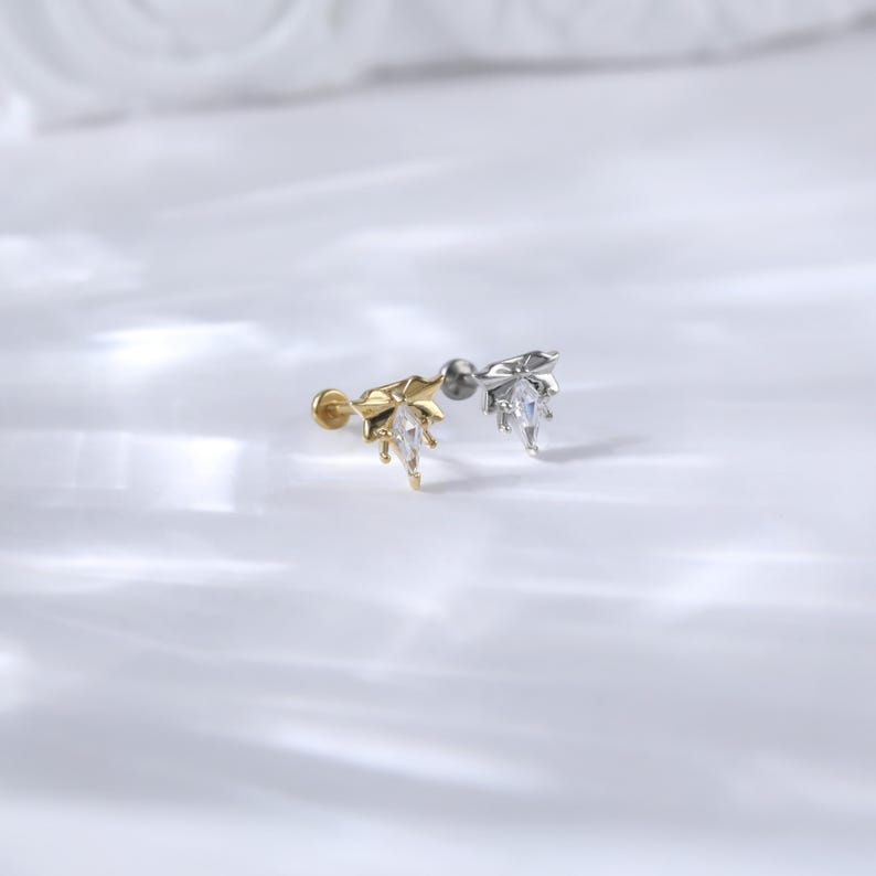 Puede incluir: Dos delicados pendientes de oreja, uno dorado y otro plateado, cada uno con un dise&ntilde;o en forma de hoja con una gema transparente en forma de l&aacute;grima. Los pendientes se presentan sobre un fondo blanco suave, resaltando sus detalles.