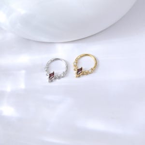 May include: Two nose rings, one silver and one gold, are displayed. Each ring features a central, dark red, diamond-shaped gem and smaller clear gems. The rings are set against a white background.